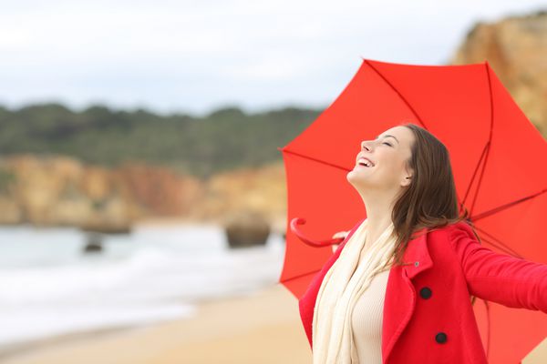 زن شاد با پوشیدن کت قرمز تنفس هوای تازه هیجان زده با یک چتر در ساحل