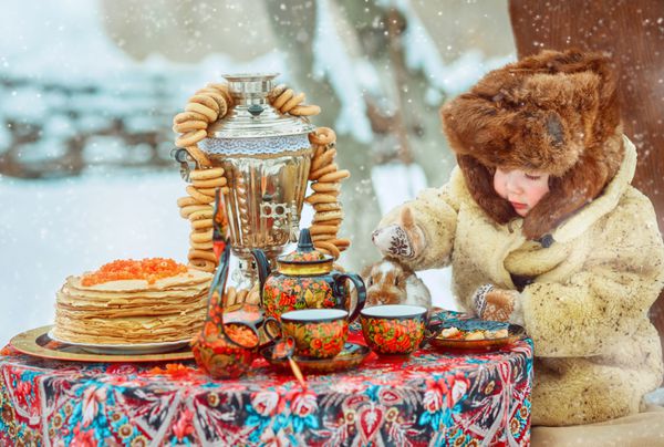 پسر کوچک در خیابان در یک کلاه خز و کت عطر و طعم Retro ملی روسیه میز برای جشن های تزئین شده است هفته پنکیک جشن آخر زمستان