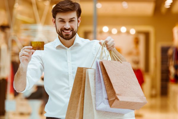 مرد جوان خوش تیپ در سفید پیراهن لبخند نگه داشتن کیف های خرید و یک کارت اعتباری در حالی که ایستاده در بازار