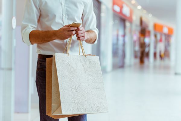 مرد جوان خوش تیپ در پیراهن سفید کیف های خرید و با استفاده از تلفن های هوشمند در حالی که ایستاده در بازار نزدیک است