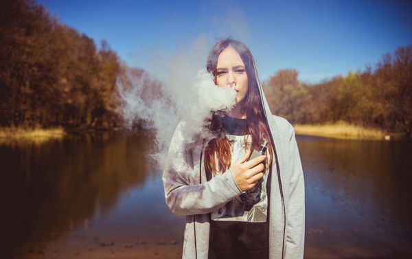 دختر ناز با یک سیگار الکترونیک در نزدیکی دریاچه پس زمینه چشم انداز