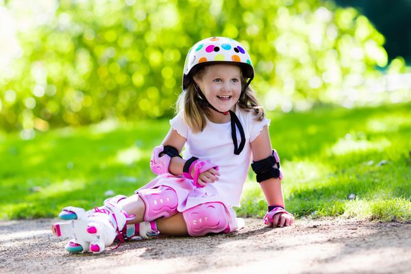 دختر کوچک یادگیری اسکیت غلتکی در پارک تابستانی آفتابی کودک پوشیدن کمربند و زانو محافظ مچ دست و کلاه ایمنی برای سوار کردن اسکیت روی غلتک ورزش در فضای باز برای بچه ها