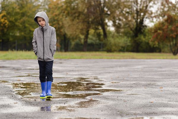 پسر کوچک ناراحت در چکمه های بارانی بارانی که در حال باریدن است روز پاییز بارانی