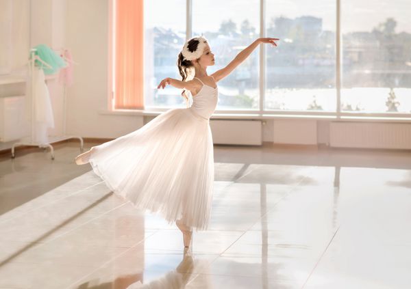 Ballerina کمی ناز در لباس های باله سفید و کفش پوینت در اتاق است بچه در کلاس دختر بچه در حال مطالعه باله است فضای کپی کنید