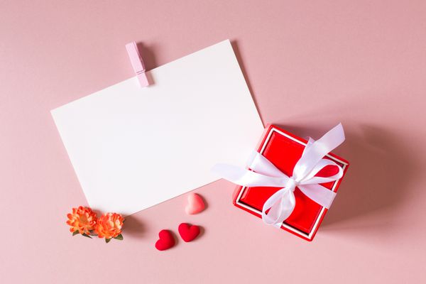 ترکیب روز جعبه هدیه قرمز با تعظیم لوازم التحریر قالب عکس با گیره قلب کوچک و گل های بهار در پس زمینه نور صورتی نمای بالا