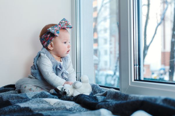 کودک کوچک با چشم آبی نشسته روی فرش در مقابل یک پنجره در زمستان
