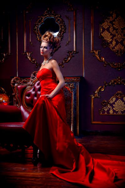 زن جوان جادویی در لباس قرمز لوکس و جواهرات گرانبها در یک صندلی در یک آپارتمان لوکس نشسته است داخلی کلاسیک زیبایی مد