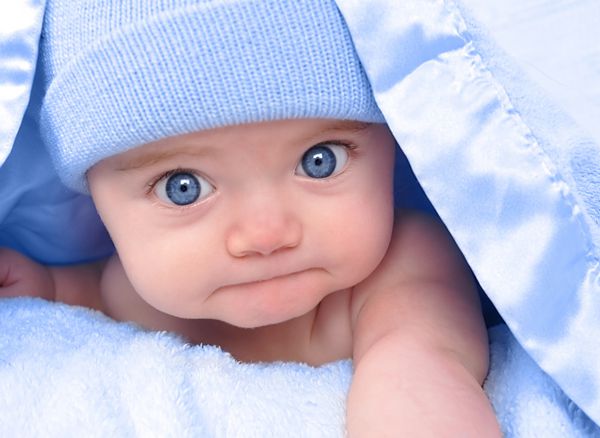 یک پسر بچه کوچک زیر پتو آبی با کلاه پنهان شده است او در دوربین خیره شده و روی شکمش ایستاده است از آن برای کودکی پدر و مادر یا مفهوم عشق استفاده کنید