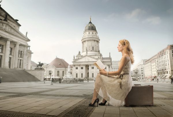 زن زیبا نشسته در چمدان و خواندن یک کتاب با بنای تاریخی در پس زمینه