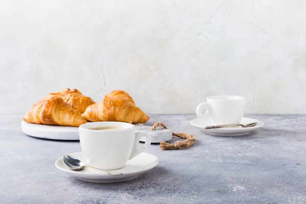 فنجان سفید قهوه و کریاسانت در پس زمینه خاکستری روشن تمرکز انتخابی مفهوم صبحانه سالم با فضای کپی