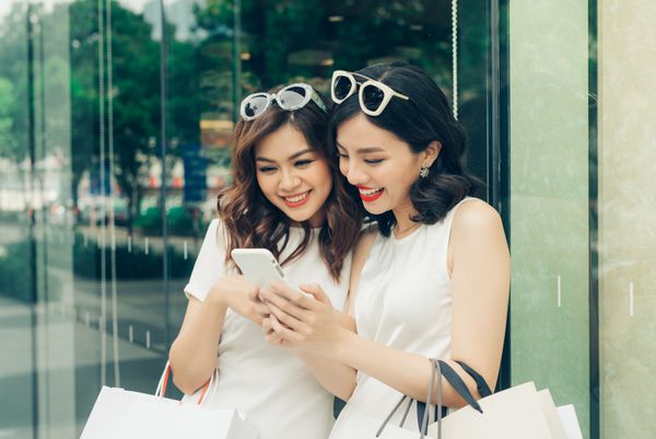 دختران زیبا آسیایی با کیسه های خرید با استفاده از گوشی های هوشمند در بازار