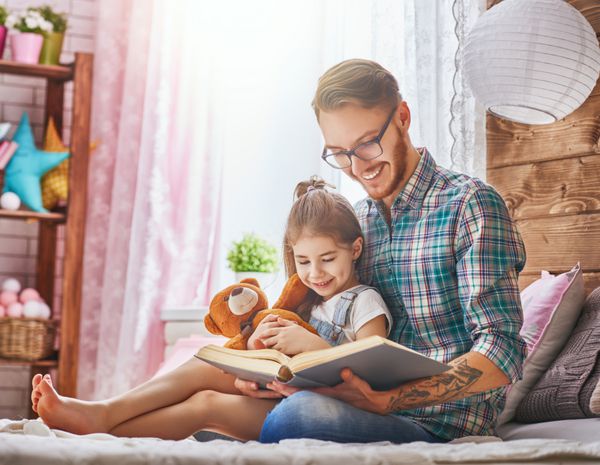 پدر و کودک روی تخت در اتاق بچه ها بابا جوان خوش تیپ خواندن یک کتاب به دخترش تعطیلات خانوادگی و همدلی