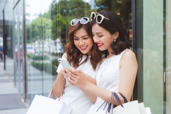 دوستان زن آسیایی خوش شانس با کیسه های با خرید لبخند زدن در حالی که نگاه کردن به تلفن در مرکز خرید