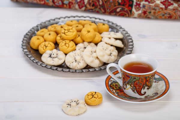 شیرینی های سنتی ایرانی گردو شکلات شیرینی کوکی نخود و کوکی های برنجی در صفحه توریوتیک فارسی با یک فنجان چاقوی فنجان طراحی پازلی و چای بشقاب بر روی زمینه سفید چوبی