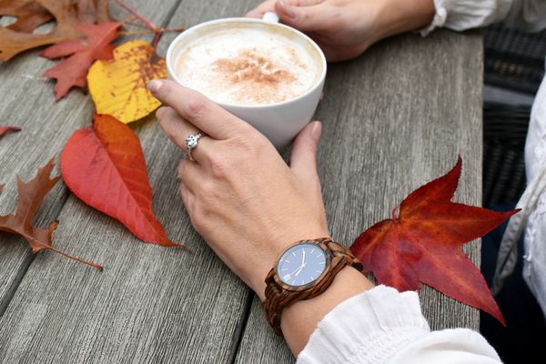سقوط زمان زن مدرن پوشیدن ساعت و نگه داشتن یک فنجان قهوه در یک میز چوبی روستایی با برگ های رنگارنگ پاییز