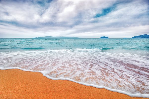 ساحل شن و ماسه آفتابی با فوم موج در آب دریا فیروزه ای و زمینه حماسی آسمان ابری یونان