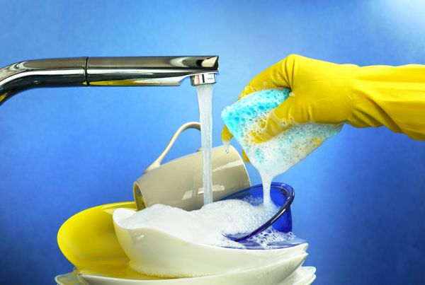 ظرف شستشو در آشپزخانه در پس زمینه آبی نزدیک کوهی از ظروف رنگی زیر شیر آب جت از شیر و دست در یک دستکش لاستیکی فشار فوم از اسفنج