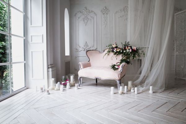 مبل مجلسی از رنگ صورتی نرم تزئین شده با گل و سبز در یک اتاق کلاسیک بر روی یک طبقه چوبی سفید قرار گرفته است که توسط شمع های روشن شده در شمع های شیشه ای در نزدیکی پنجره بزرگ و پرده ها قرار دارد