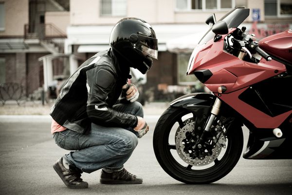 یک مرد موتورسیکلت در یک کلاه ایمنی و یک کاپشن چرم و شلوار جین مخالف یک موتورسیکلت از یک رنگ قرمز ورزشی است و به او نگاه می کند به صورت