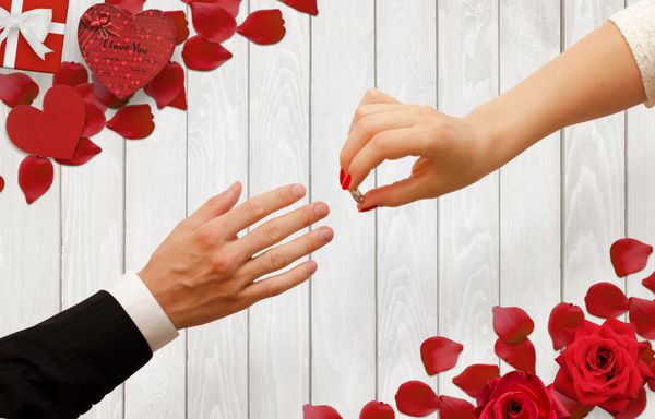 زن حلقه نامزدی را در دست انسان قرار دادن پس زمینه رمانتیک با گلبرگ گل سرخ هدیه بر روی میز