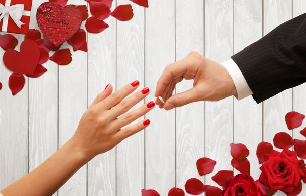 قرار دادن حلقه نامزدی را در دست زن مرد پس زمینه رمانتیک با گلبرگ گل سرخ هدیه بر روی میز