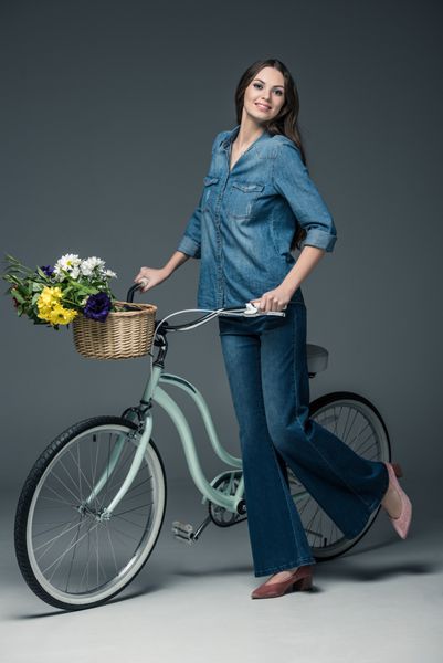 دختر زیبا در جین لباس نگه داشتن دوچرخه با گل در سبد ترکه در خاکستری