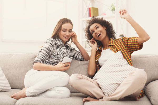 دو زن گوش دادن به موسیقی آنلاین بر روی گوشی های هوشمند و به اشتراک گذاری گوشی های نشسته در نیمکت در خانه فضای کپی