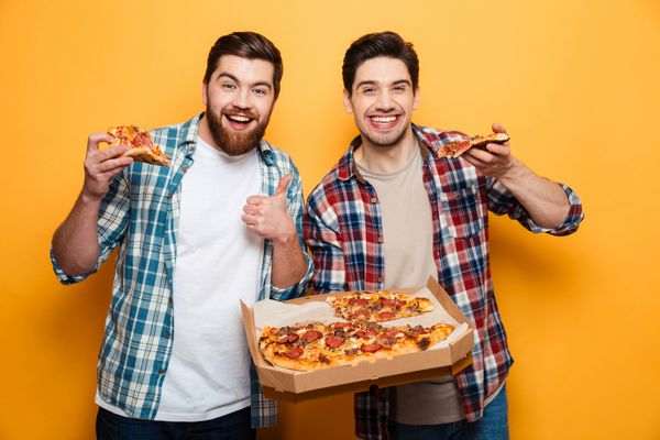 دو مرد شاد که پیتزا را نگه داشته و به دوربین نگاه می کند در حالی که مرد ریش دار انگشت شستی بیش از پس زمینه ی زرد دارد
