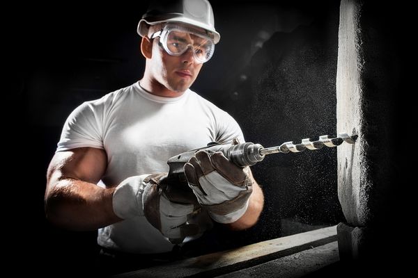 مرد با یک ابزار کارگر با استفاده از ابزار قدرت حفاری در محل ساخت و ساز مرد جوان با استفاده از برق بر روی دیوار آجری سفید در کلاه سفید