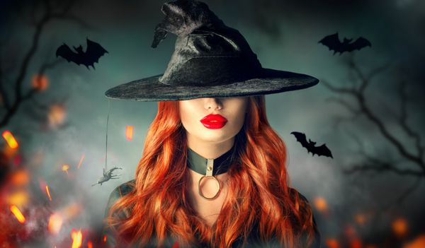 عکس دختر جادوگر هالووین زن زیبا زن جوان در جادوگران کلاه با موی قرمز موی قرمز و آرایش لب روشن بیش از حوادث پس زمینه جنگل سحر و جادو تاریک تاریک طراحی هنری حزب هالووین