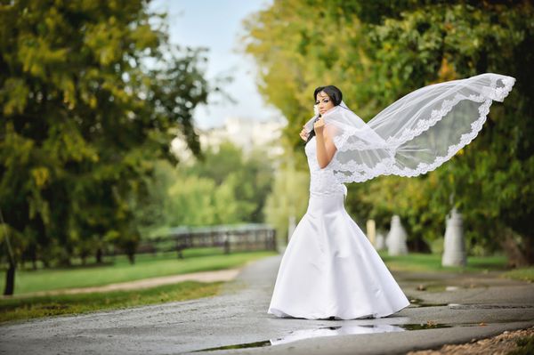 در روز عروسی خود عروس جوان دوست داشتنی در وسط جاده در پارک در طبیعت با حجاب جریان می ایستد