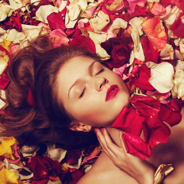 پرتره شیک مدل با موهای قرمز زنجبیل با لب های قرمز دراز کشیدن بر روی گلبرگ رز زمینه محو شدن استودیو شات