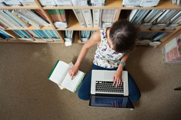 دیدگاه بالا از یک دانش آموز دختر نشسته در برابر قفسه کتاب با لپ تاپ در طبقه کتابخانه