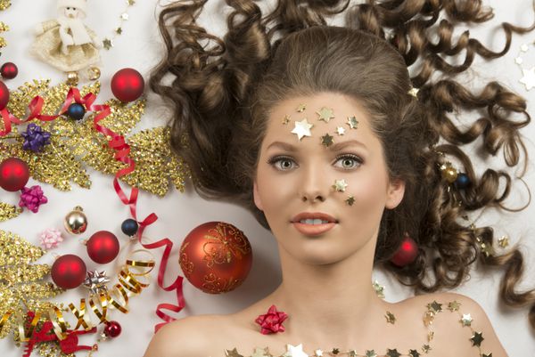 پرتره از دختر کریسمس پر زرق و برق با موهای فرفری طولانی قهوه ای و ستاره های آرایش صورت با دکوراسیون های مختلف کریسمس بی رنگ قرمز و رنگارنگ روبان ستاره های طلایی و شاخه