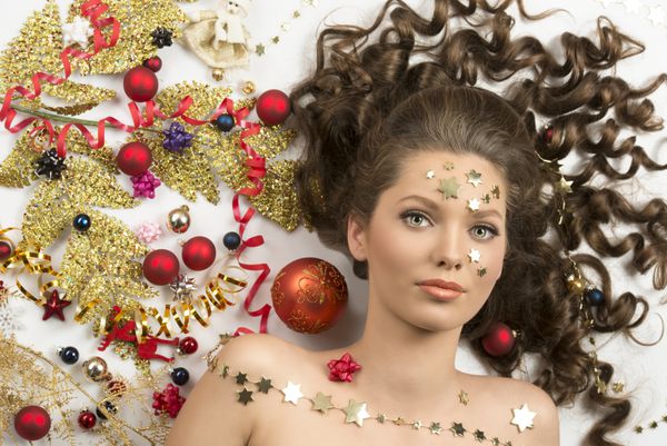 زن جذاب با موهای فرفره ای که در نزدیکی زیبایی پرتره احاطه شده توسط برخی از دکوراسیون کریسمس بطری های سرخ شاخه طلایی روبان و ستاره ها