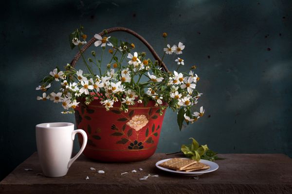 سبد قرمز با گل های سفید فنجان قهوه و بیسکویت روی میز چوبی
