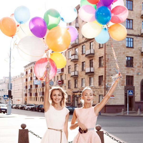 دو دختر خندان در لباس های سفید و صورتی که شمشیرهای بالن های رنگارنگ در خورشید در خیابان شهر دارند