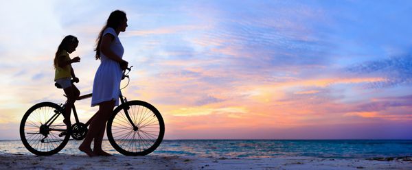پانورامای مادر و دختر با دوچرخه سواری در یک ساحل در غروب آفتاب