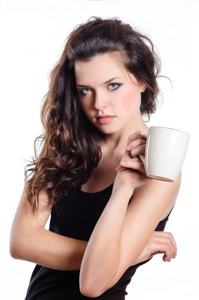 زن جوان با موهای تیره نگاه کردن به دوربین نگه داشتن فنجان قهوه یا چای در دست او جدا شده بر روی زمینه سفید