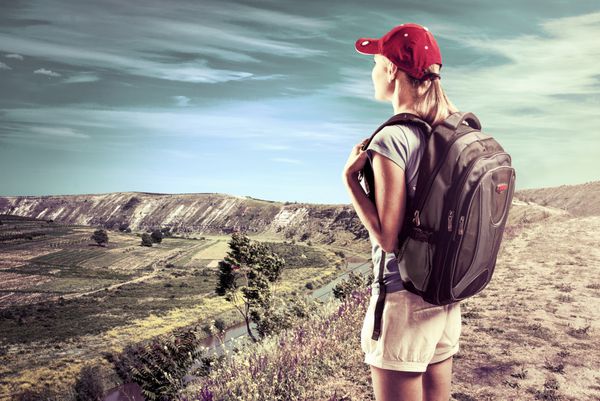 کوهنورد زن به منظره کوهستانی زیبا نگاه می کند جوان گردشگر زن فعال در بالای تپه با کوله پشتی که طبیعت و آسمان آبی را تحسین می کند ایستاده است