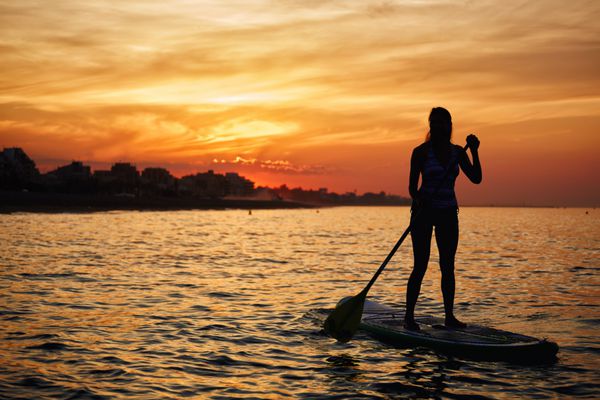 شبح خیره کننده از یک دختر زیبا شناور در تخته موج سواری در غروب آفتاب نارنجی شگفت انگیز بیش از دریا