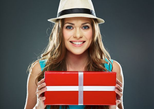 زن جوان با لبخند دندان بزرگ جعبه هدیه قرمز را در برابر پس زمینه خاکستری نگه دارید