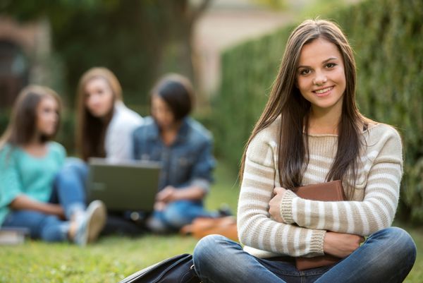 پرتره از یک دختر کالج خندان در حال برگزاری کتاب با دانشجویان مبهم است که در پارک نشسته اند