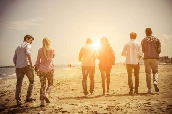 گروه چند قومی از دوستان پیاده روی در ساحل و صحبت کردن گروه جوانان شکلات در غروب آفتاب