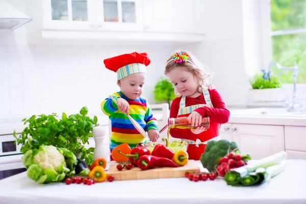بچه ها سالاد سبزیجات تازه را در یک آشپزخانه سفید آشپزی می کنند کودکان سبزیجات را برای ناهار گیاهی آماده می کنند کودک نوپا و کودک شام صبحانه بخورند پسر و دختر آماده و خوردن وعده غذایی خام است تغذیه کودک