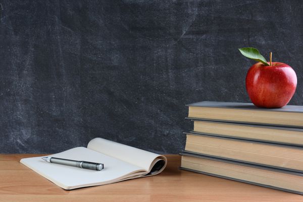 نزدیک یک میز معلم با کتاب کاغذ و قلم و سیب قرمز در مقابل تخته سیاه فرمت افقی با فضای کپی