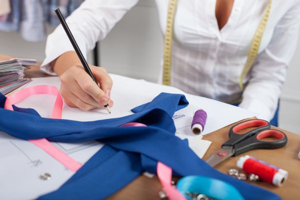 نزدیک دست از طراح مد لباس ماهر زن نشسته روی میز است او نقاشی لباس را بر روی یک طرح طراحی کرده است چیزهای طراحی زیادی روی میز وجود دارد