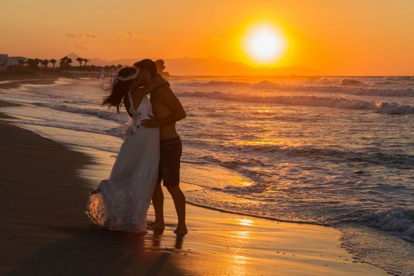 با نور پس زمینه به تازگی زن و شوهر جوان در ساحل شکل گرفته لذت بردن از غروب خفیف غلیظ پوشیدن لباس عروسی و شورت پابرهنه مرطوب اذیت کردن و بوسیدن یکدیگر
