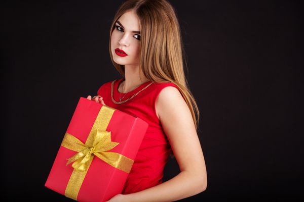 دختر زیبا پوشیدن لباس قرمز نگه داشتن جعبه هدیه است لبخند پرتره زن زیبا بر روی زمینه سیاه و سفید جدا شده است