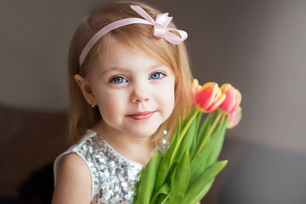 دختر کوچک با یک دسته گل لاله به رنگ در یک لباس سفید با یک روبان در موهایش تصویر افقی به دوربین نگاه کرد با یک لبخند ملایم